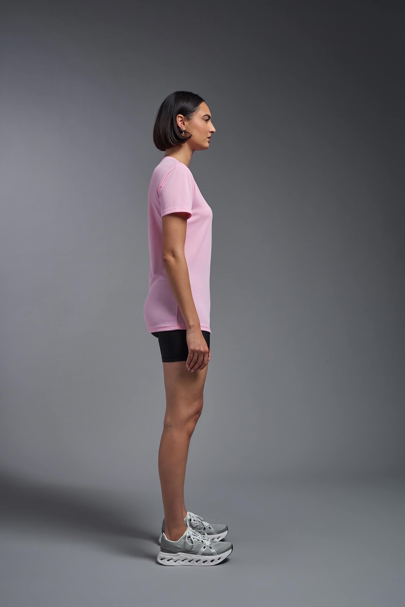 A female model with brown short hair on a gray background, wearing the Women's Oxymesh™ V-neck Tech Tee in the color pink from a full body side view. This shirt is made in the USA and is quick-drying and moisture-wicking. #color_pink
