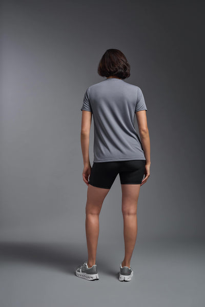 A female model with brown short hair on a gray background, wearing the Women's Oxymesh™ V-neck Tech Tee in the color steel gray from a full body back view. This shirt is made in the USA and is quick-drying and moisture-wicking. #color_steel-gray