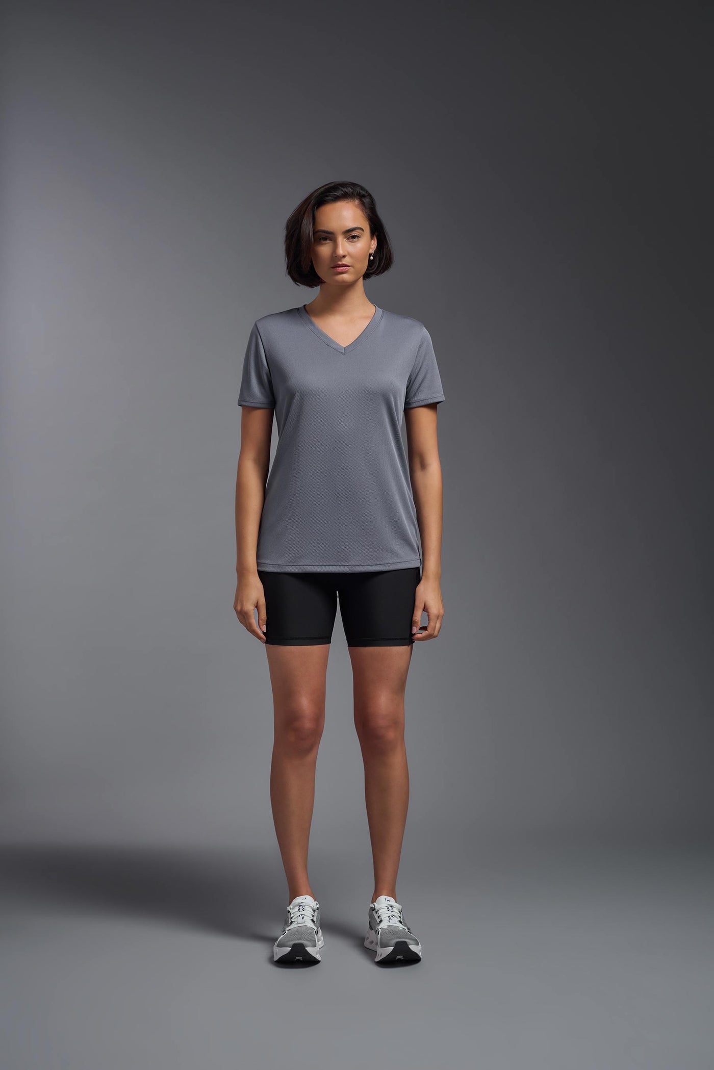 A female model with brown short hair on a gray background, wearing the Women's Oxymesh™ V-neck Tech Tee in the color steel gray from a full body front view. This shirt is made in the USA and is quick-drying and moisture-wicking. #color_steel-gray