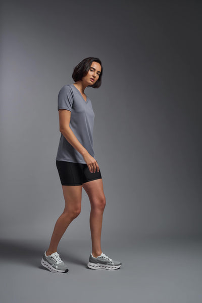 A female model with brown short hair on a gray background, wearing the Women's Oxymesh™ V-neck Tech Tee in the color steel gray from a full body side view. This shirt is made in the USA and is quick-drying and moisture-wicking. #color_steel-gray