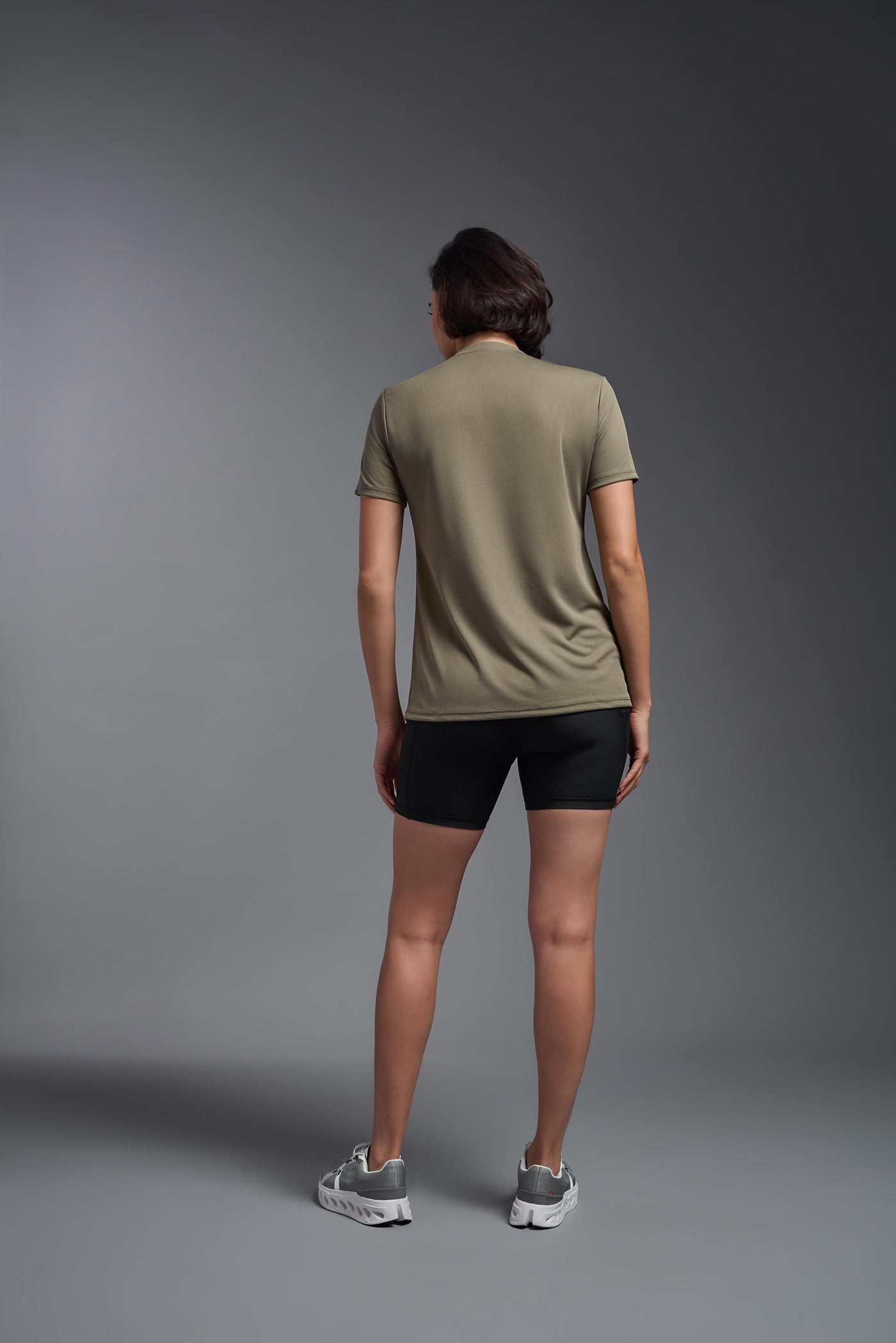 A female model with brown short hair on a gray background, wearing the Women's Oxymesh™ V-neck Tech Tee in the color tan-499 from a full body back view. This shirt is made in the USA and is quick-drying and moisture-wicking. #color_tan-499