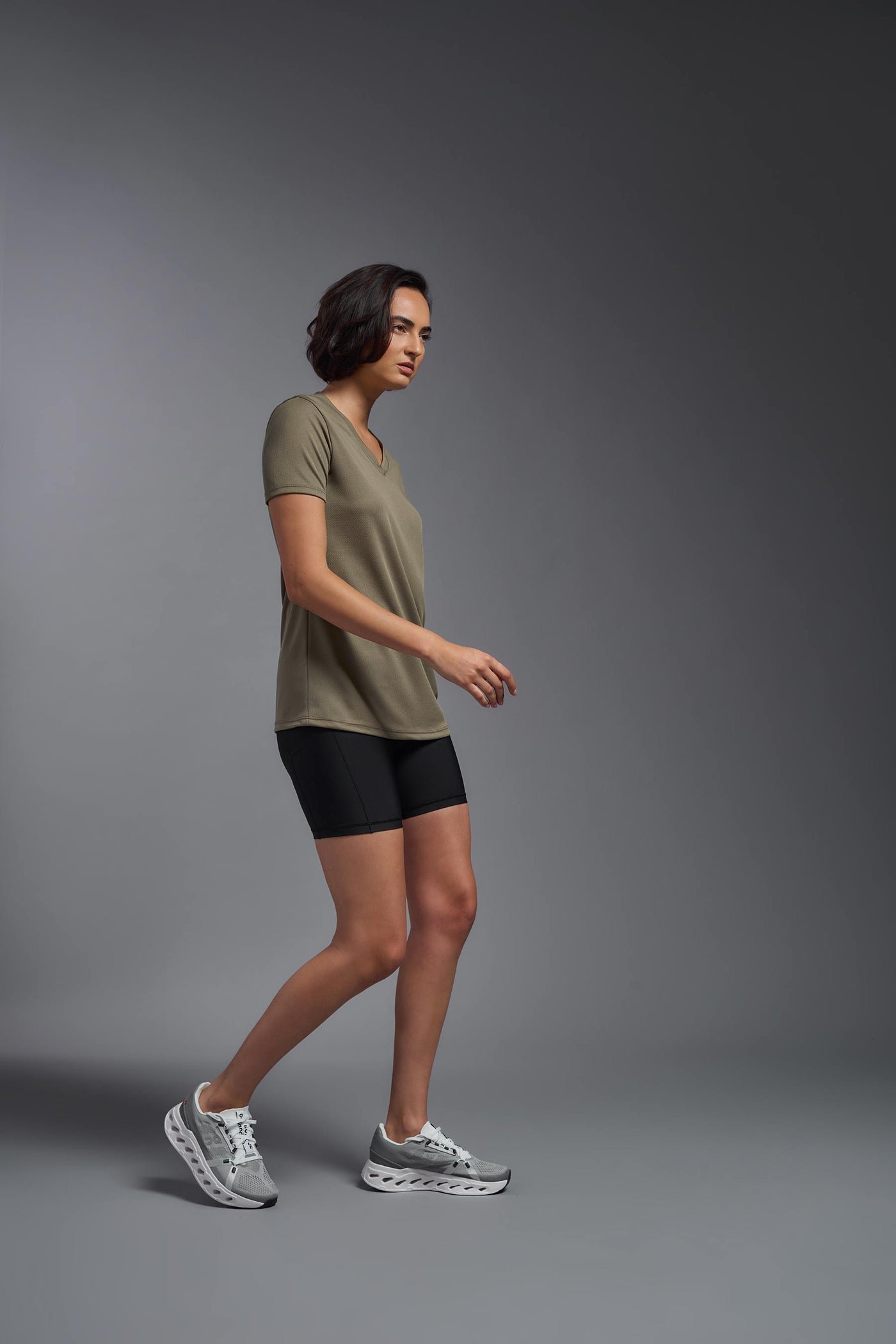 A female model with brown short hair on a gray background, wearing the Women's Oxymesh™ V-neck Tech Tee in the color tan-499 from a full body side view. This shirt is made in the USA and is quick-drying and moisture-wicking. #color_tan-499