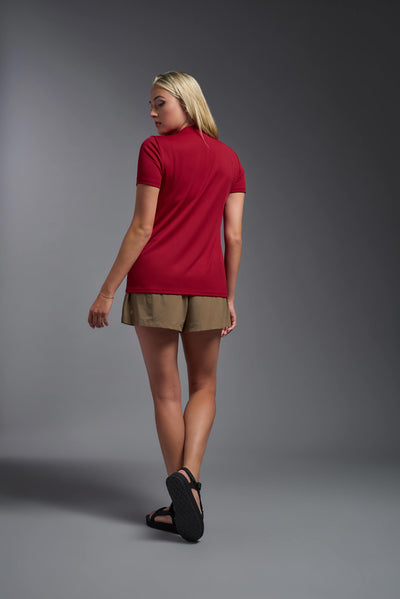 A blonde female model with long straight hair on a gray background, wearing the Women's Oxymesh™ V-neck Tech Tee in the color true red from a full body back view. This shirt is made in the USA and is quick-drying and moisture-wicking. #color_true-red
