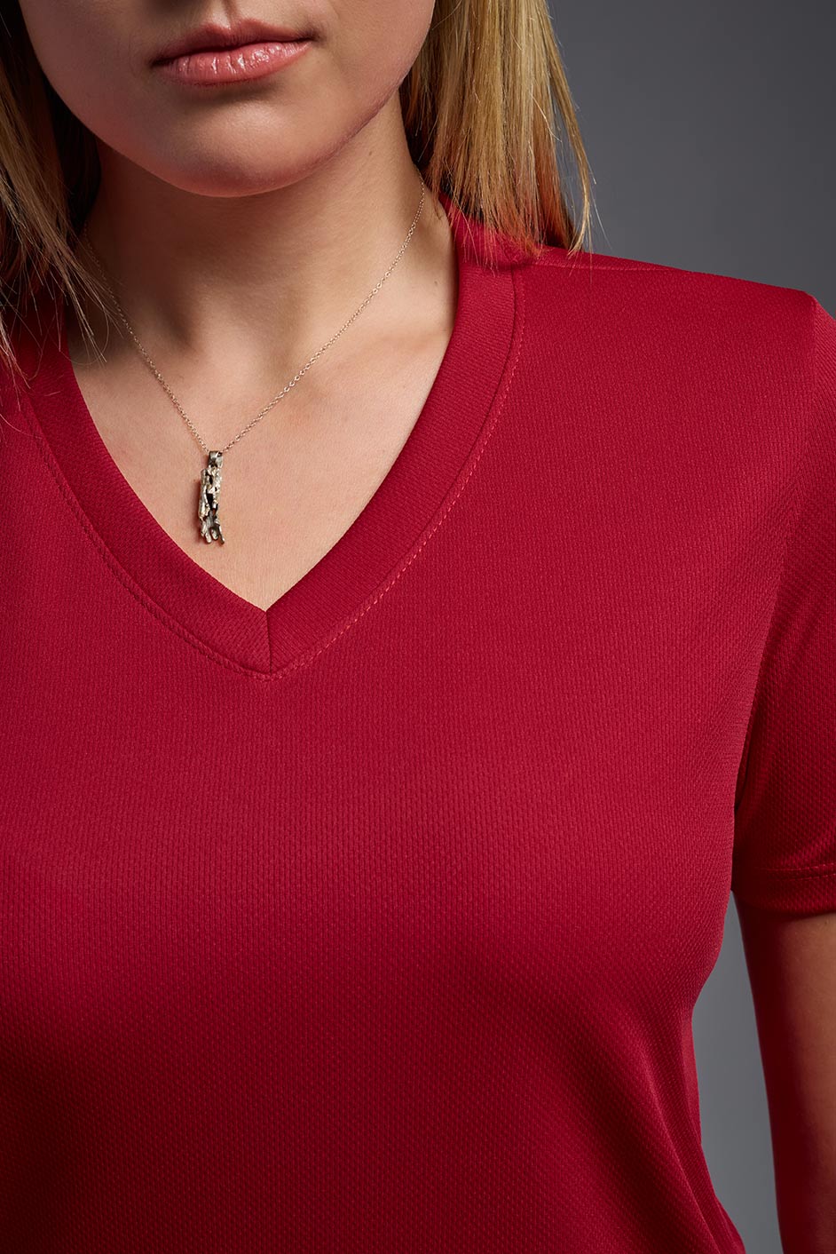 A blonde female model with long straight hair on a gray background, wearing the Women's Oxymesh™ V-neck Tech Tee in the color true red from a closeup collar front view. This shirt is made in the USA and is quick-drying and moisture-wicking. #color_true-red