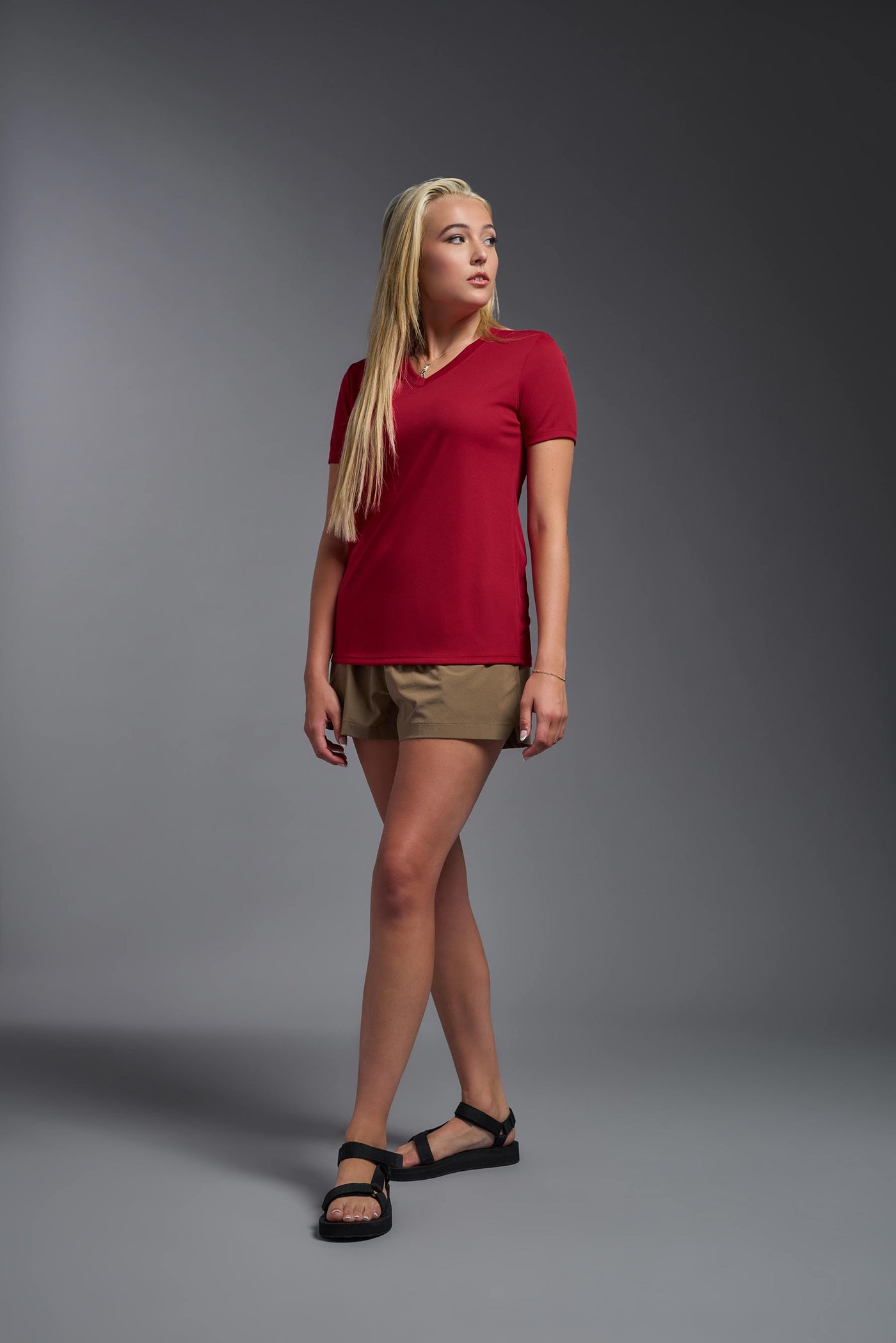 A blonde female model with long straight hair on a gray background, wearing the Women's Oxymesh™ V-neck Tech Tee in the color true red from a full body side view. This shirt is made in the USA and is quick-drying and moisture-wicking. #color_true-red