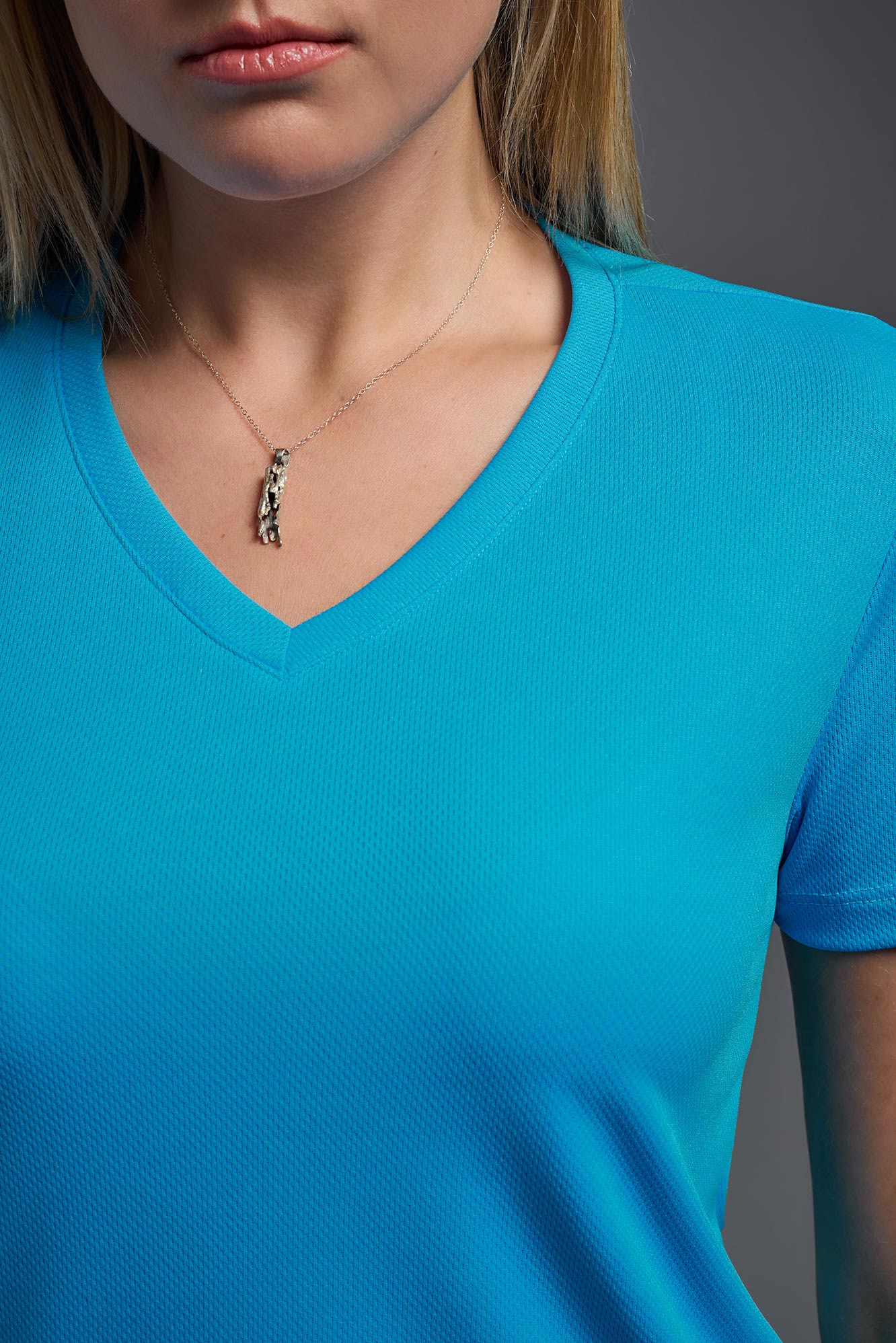 A blonde female model with long straight hair on a gray background, wearing the Women's Oxymesh™ V-neck Tech Tee in the color turquoise from a closeup collar front view. This shirt is made in the USA and is quick-drying and moisture-wicking. #color_turquoise