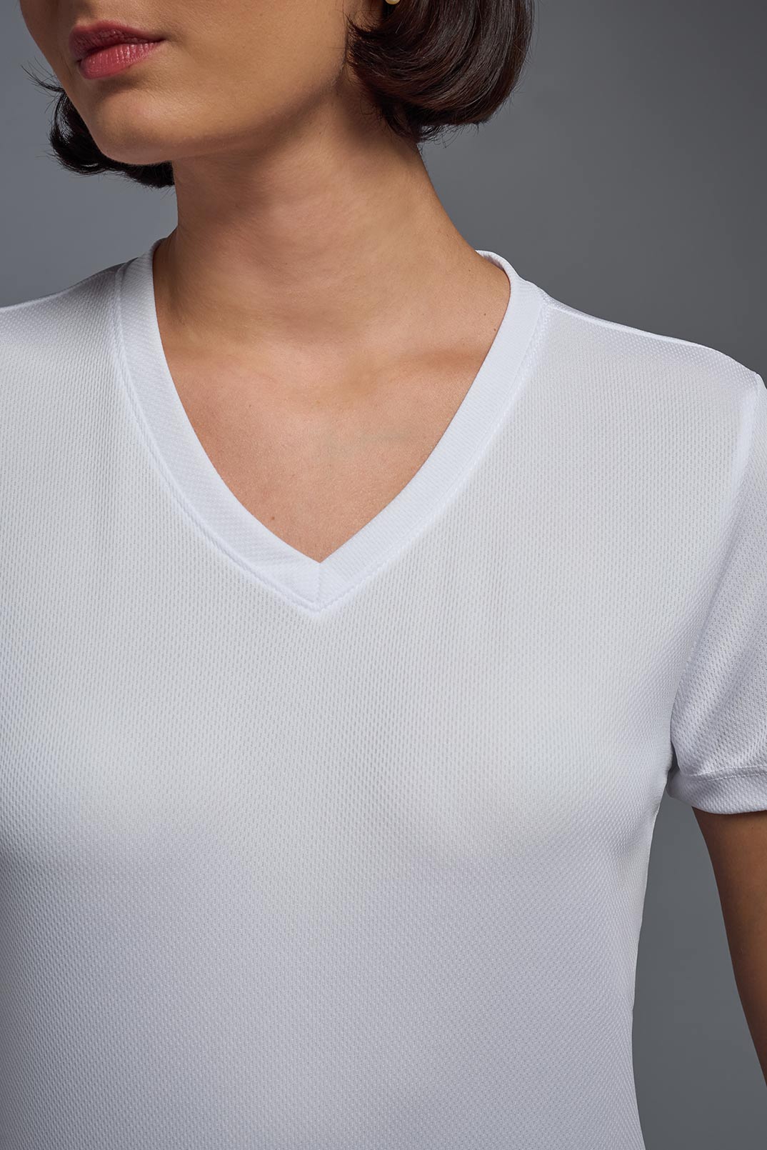 A female model with brown short hair on a gray background, wearing the Women's Oxymesh™ V-neck Tech Tee in the color white from a closeup collar front view. This shirt is made in the USA and is quick-drying and moisture-wicking. #color_white