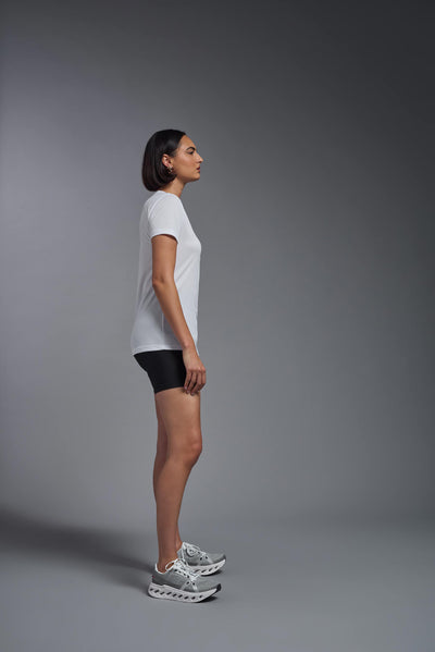 A female model with brown short hair on a gray background, wearing the Women's Oxymesh™ V-neck Tech Tee in the color white from a full body side view. This shirt is made in the USA and is quick-drying and moisture-wicking. #color_white