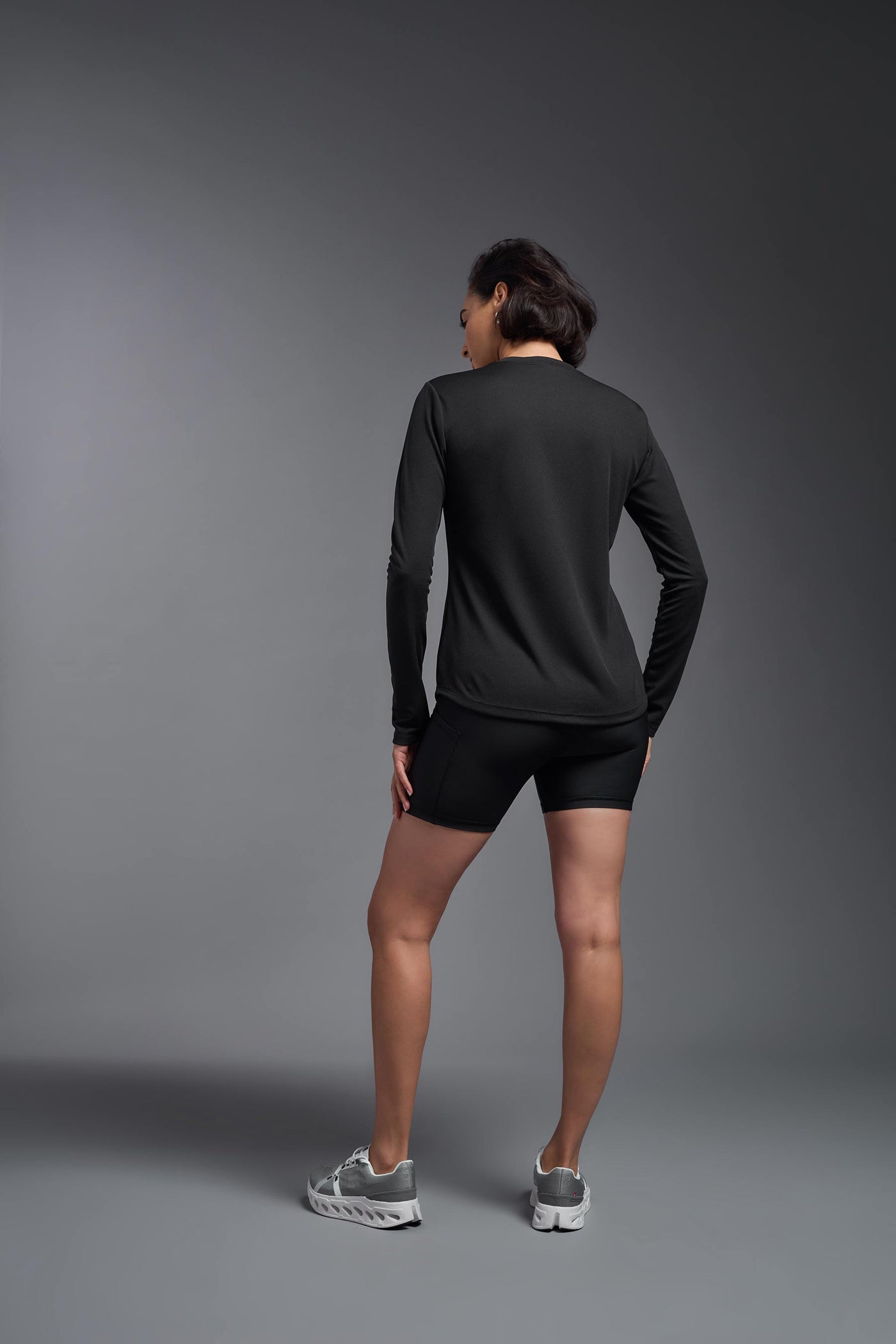 A female model with short brown hair on a gray background, wearing the Women's Oxymesh™ Long Sleeve Tech Tee in the color black from a full body back view. This shirt is made in the USA and is quick-drying and moisture-wicking. #color_black
