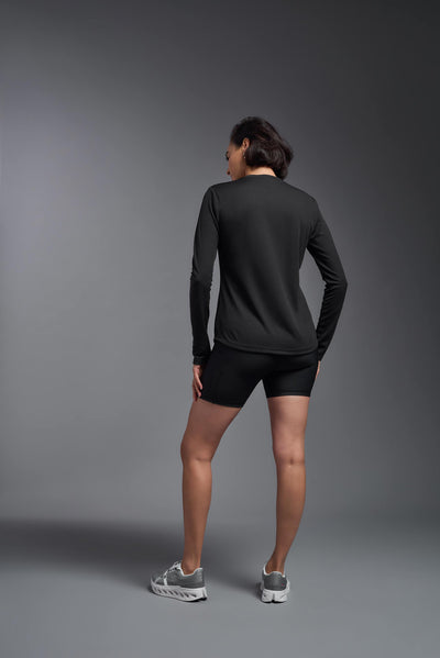 A female model with short brown hair on a gray background, wearing the Women's Oxymesh™ Long Sleeve Tech Tee in the color black from a full body back view. This shirt is made in the USA and is quick-drying and moisture-wicking. #color_black