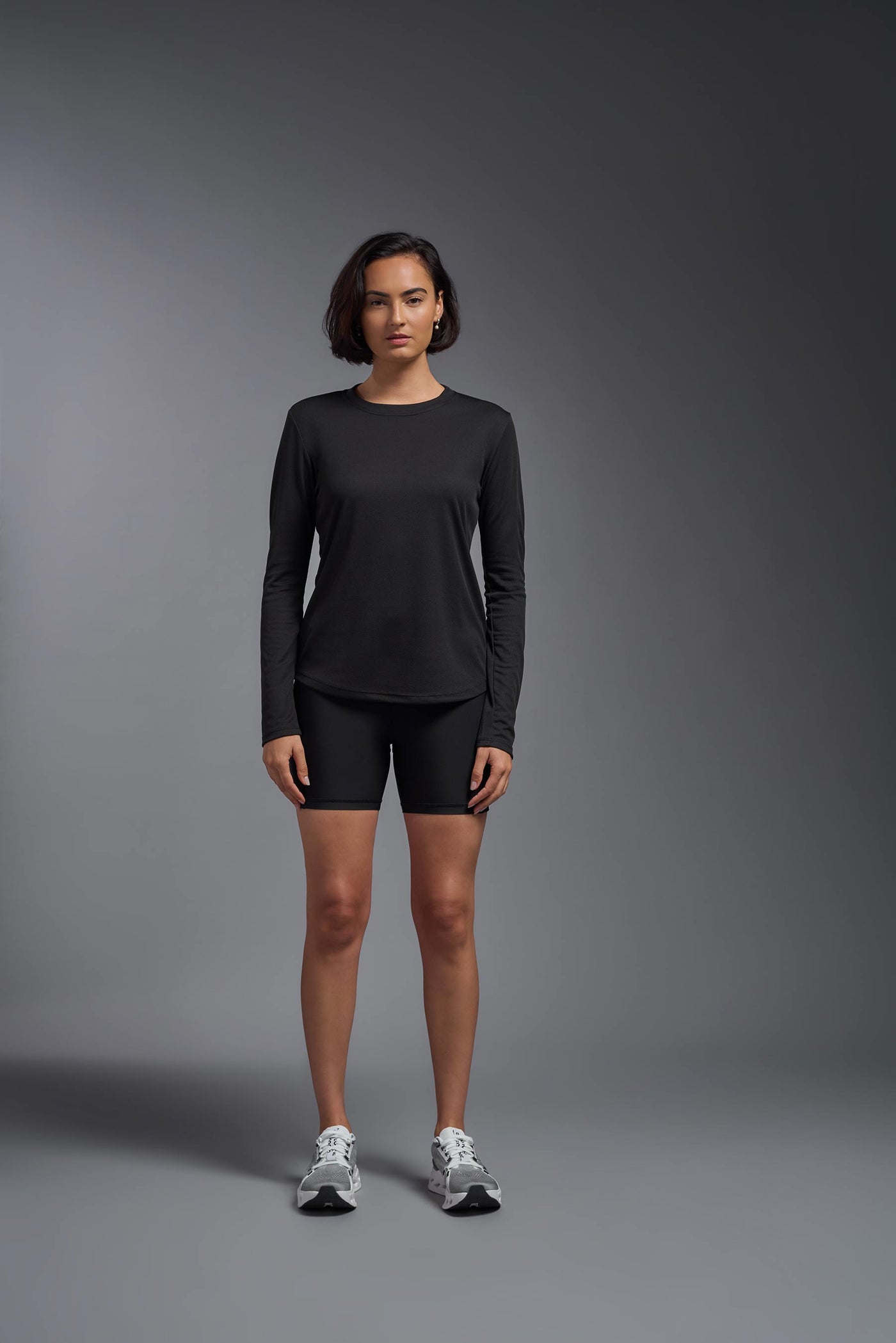A female model with short brown hair on a gray background, wearing the Women's Oxymesh™ Long Sleeve Tech Tee in the color black from a full body front view. This shirt is made in the USA and is quick-drying and moisture-wicking. #color_black
