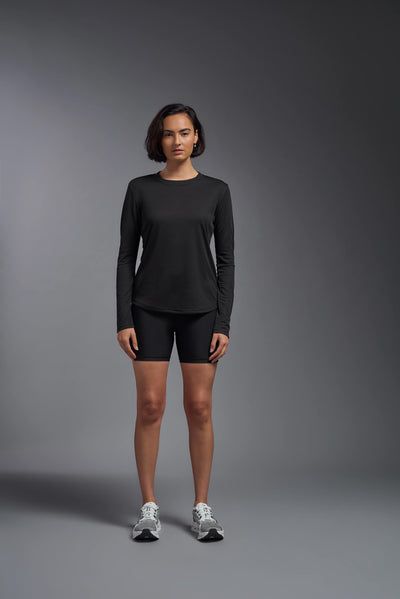 A female model with short brown hair on a gray background, wearing the Women's Oxymesh™ Long Sleeve Tech Tee in the color black from a full body front view. This shirt is made in the USA and is quick-drying and moisture-wicking. #color_black