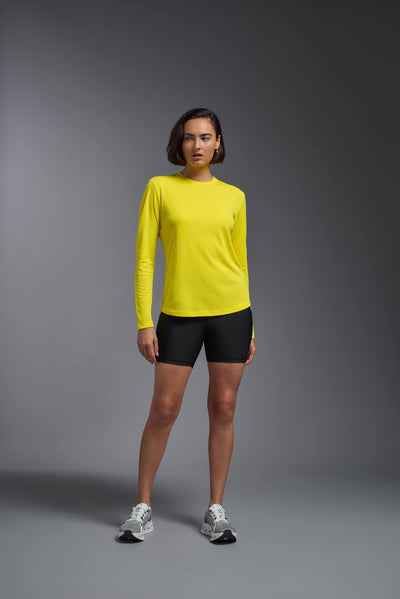 A female model with short brown hair on a gray background, wearing the Women's Oxymesh™ Long Sleeve Tech Tee in the color bright yellow from a full body front view. This shirt is made in the USA and is quick-drying and moisture-wicking. #color_bright-yellow
