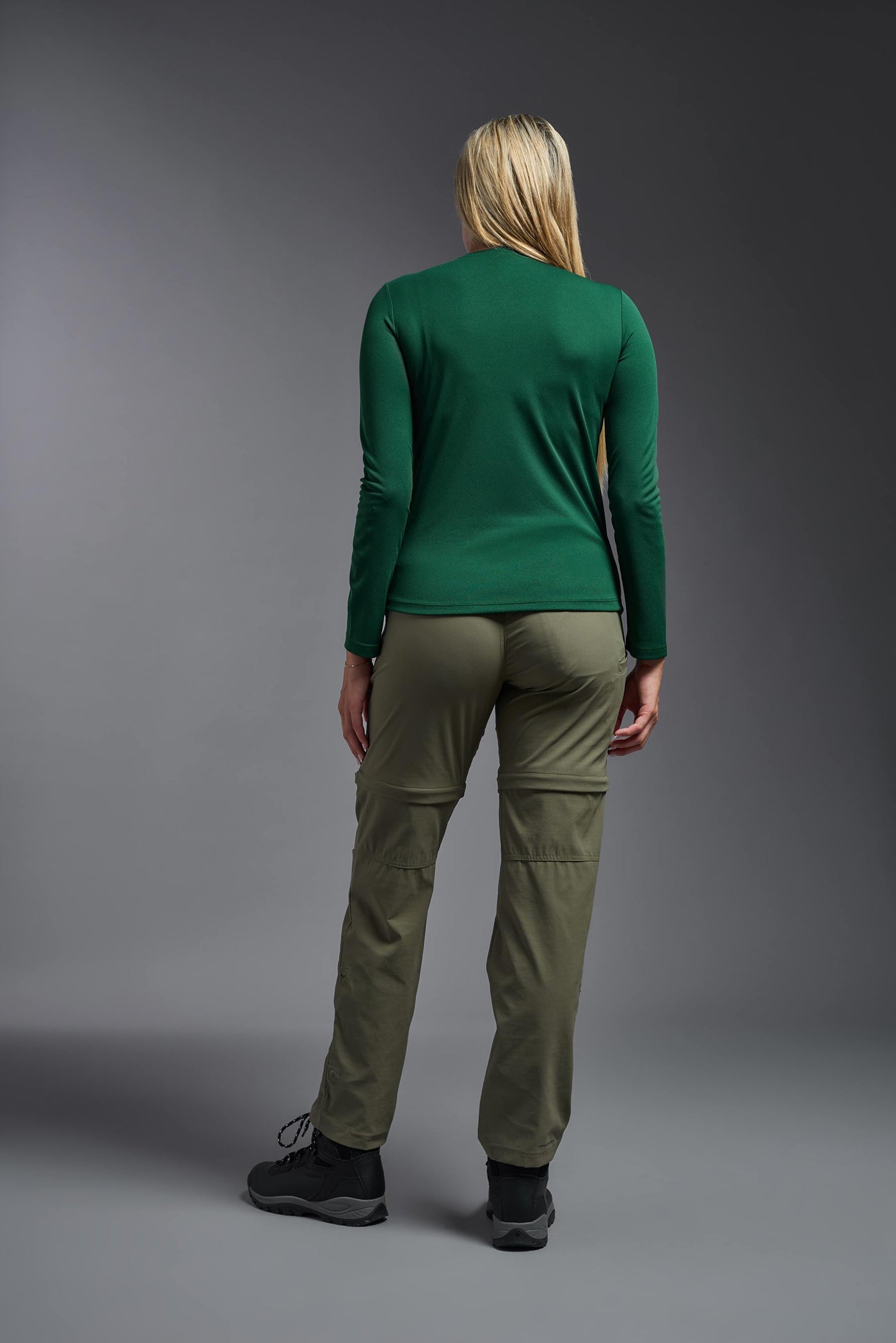 A blonde, female model with long straight hair on a gray background, wearing the Women's Oxymesh™ Long Sleeve Tech Tee in the color forest green from a full body back view. This shirt is made in the USA and is quick-drying and moisture-wicking. #color_forest-green