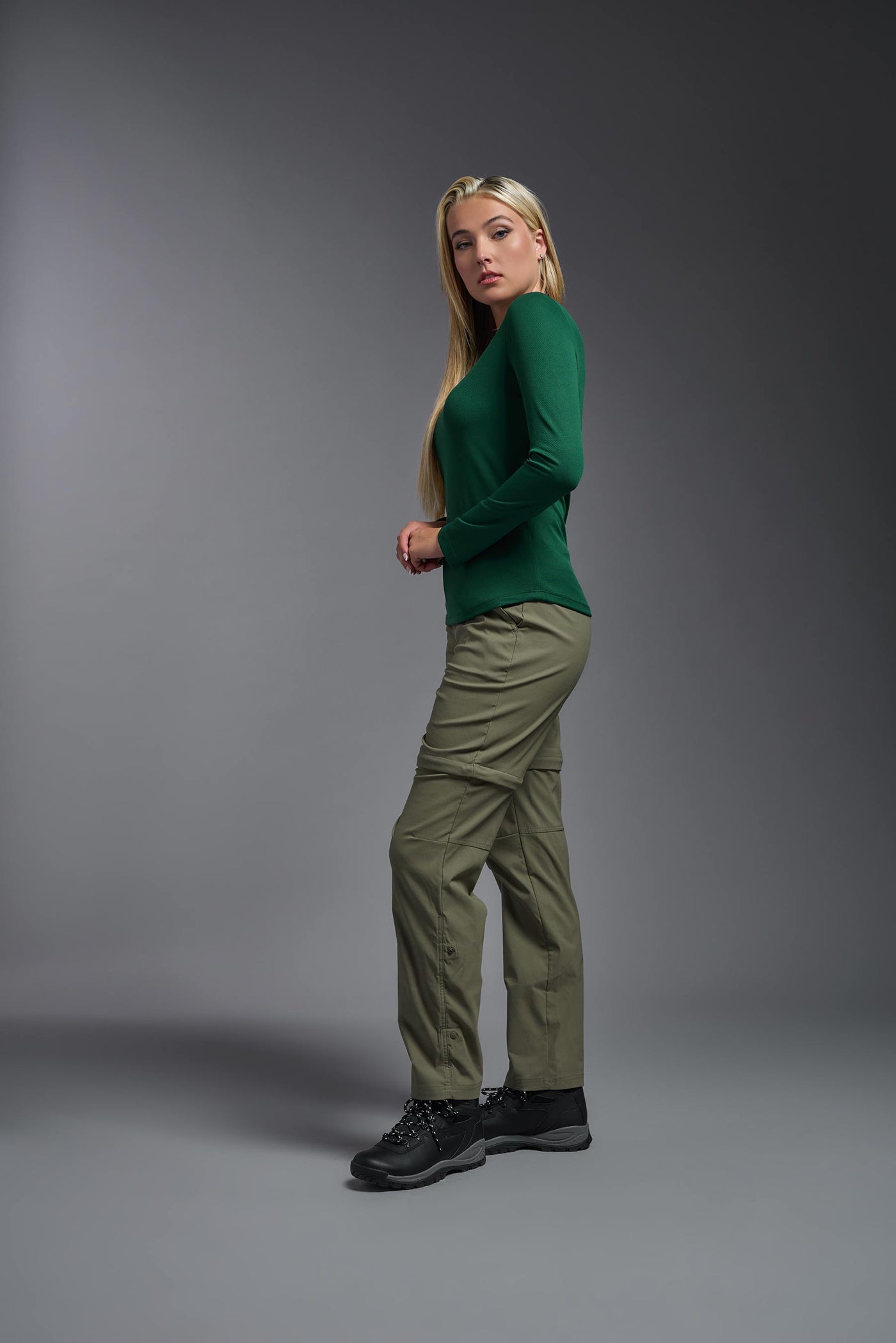 A blonde, female model with long straight hair on a gray background, wearing the Women's Oxymesh™ Long Sleeve Tech Tee in the color forest green from a full body side view. This shirt is made in the USA and is quick-drying and moisture-wicking. #color_forest-green