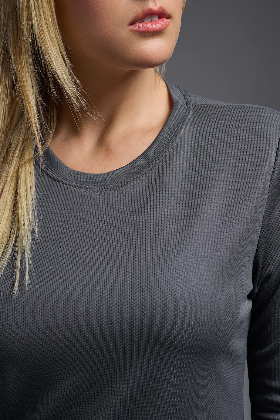 A blonde, female model with long straight hair on a gray background, wearing the Women's Oxymesh™ Long Sleeve Tech Tee in the color graphite gray from a closeup collar front view. This shirt is made in the USA and is quick-drying and moisture-wicking. #color_graphite-gray
