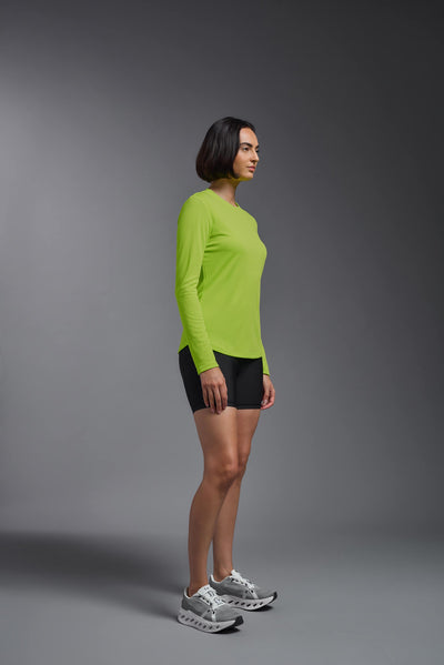 A female model with short brown hair on a gray background, wearing the Women's Oxymesh™ Long Sleeve Tech Tee in the color key lime from a full body side view. This shirt is made in the USA and is quick-drying and moisture-wicking. #color_key-lime