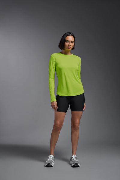 A female model with short brown hair on a gray background, wearing the Women's Oxymesh™ Long Sleeve Tech Tee in the color key lime from a full body front view. This shirt is made in the USA and is quick-drying and moisture-wicking. #color_key-lime