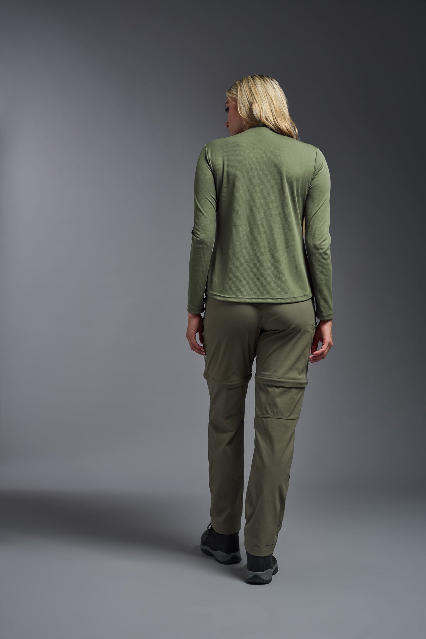 A blonde female model with long straight hair on a gray background, wearing the Women's Oxymesh™ Long Sleeve Tech Tee in the color military green from a full body back view. This shirt is made in the USA and is quick-drying and moisture-wicking. #color_military-green