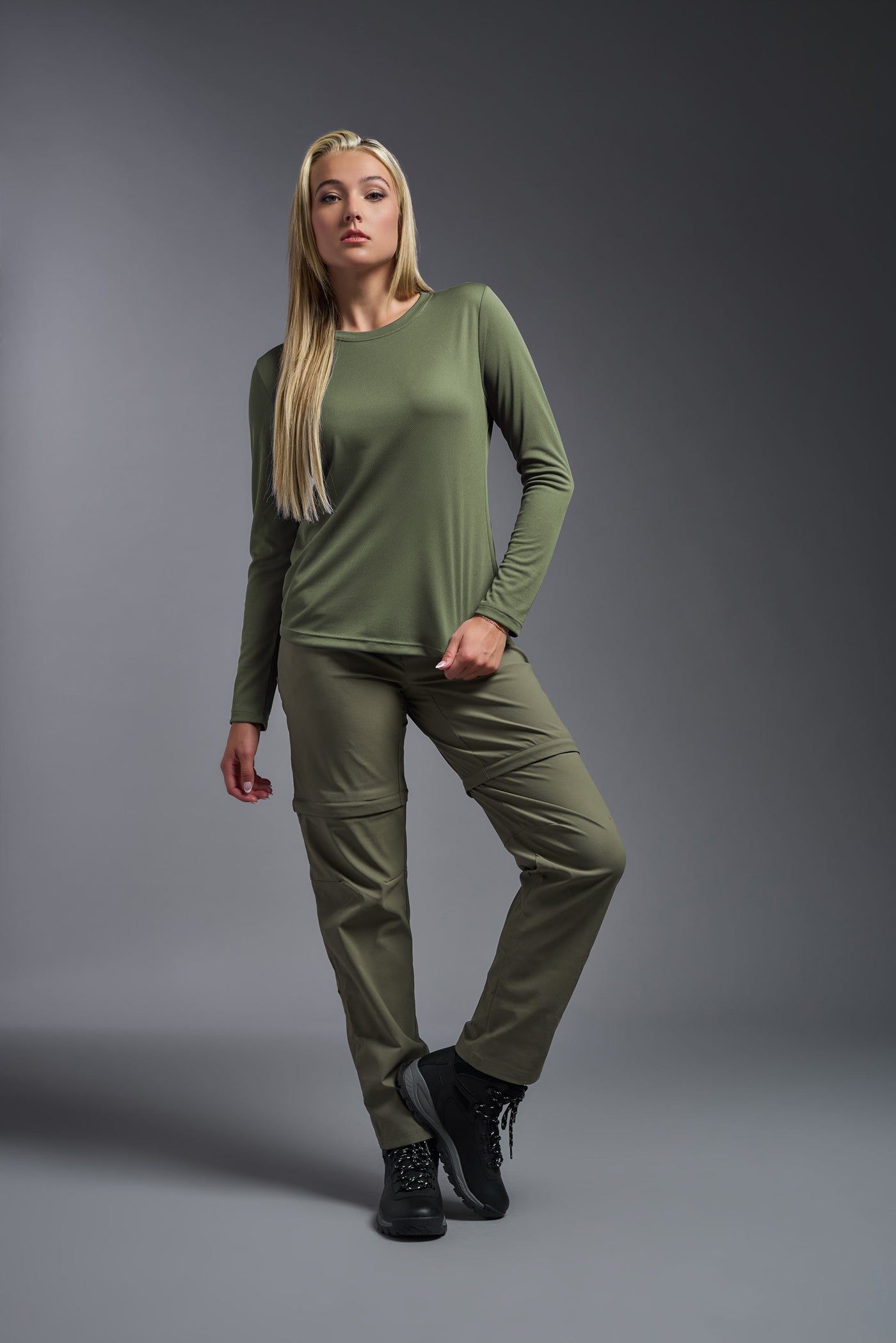 A blonde female model with long straight hair on a gray background, wearing the Women's Oxymesh™ Long Sleeve Tech Tee in the color military green from a full body front view. This shirt is made in the USA and is quick-drying and moisture-wicking. #color_military-green