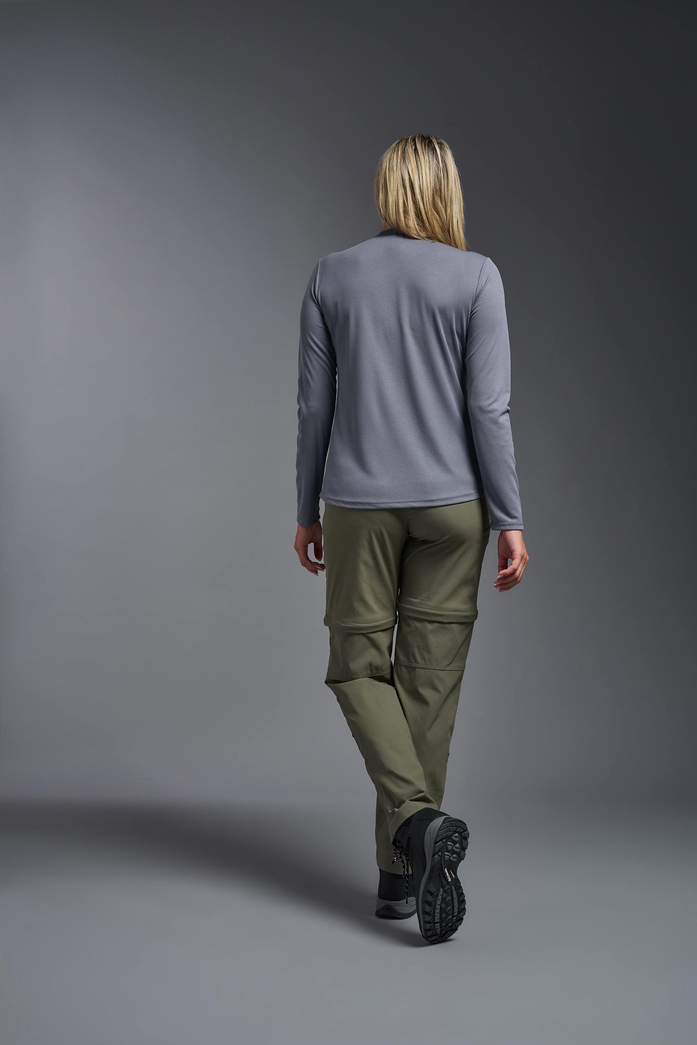 A blonde, female model with long straight hair on a gray background, wearing the Women's Oxymesh™ Long Sleeve Tech Tee in the color steel gray from a full body back view. This shirt is made in the USA and is quick-drying and moisture-wicking. #color_steel-gray