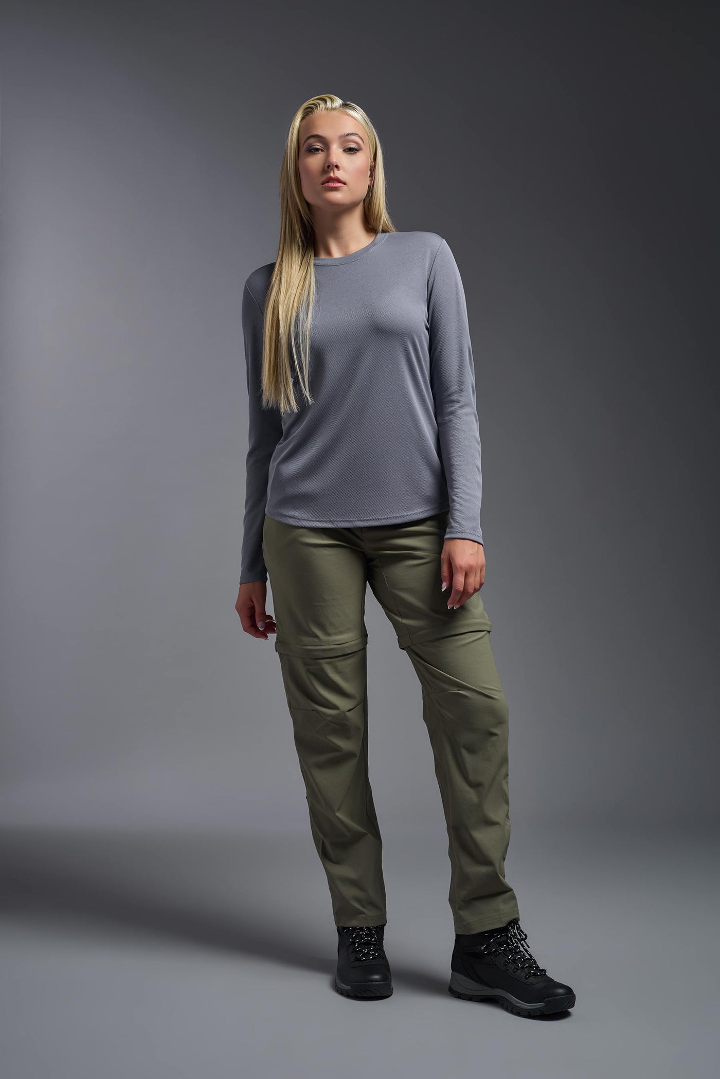A blonde, female model with long straight hair on a gray background, wearing the Women's Oxymesh™ Long Sleeve Tech Tee in the color steel gray from a full body front view. This shirt is made in the USA and is quick-drying and moisture-wicking. #color_steel-gray