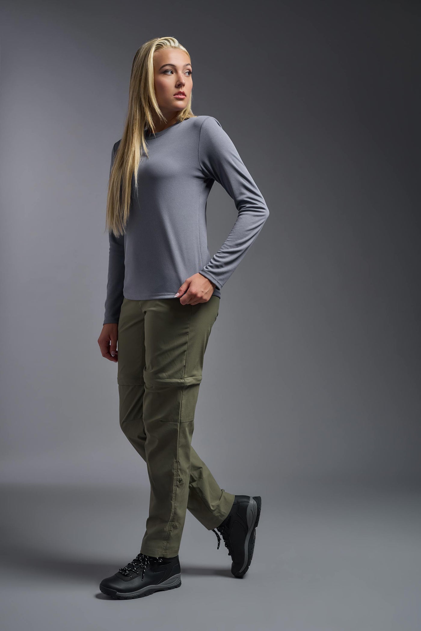 A blonde, female model with long straight hair on a gray background, wearing the Women's Oxymesh™ Long Sleeve Tech Tee in the color steel gray from a full body side view. This shirt is made in the USA and is quick-drying and moisture-wicking. #color_steel-gray