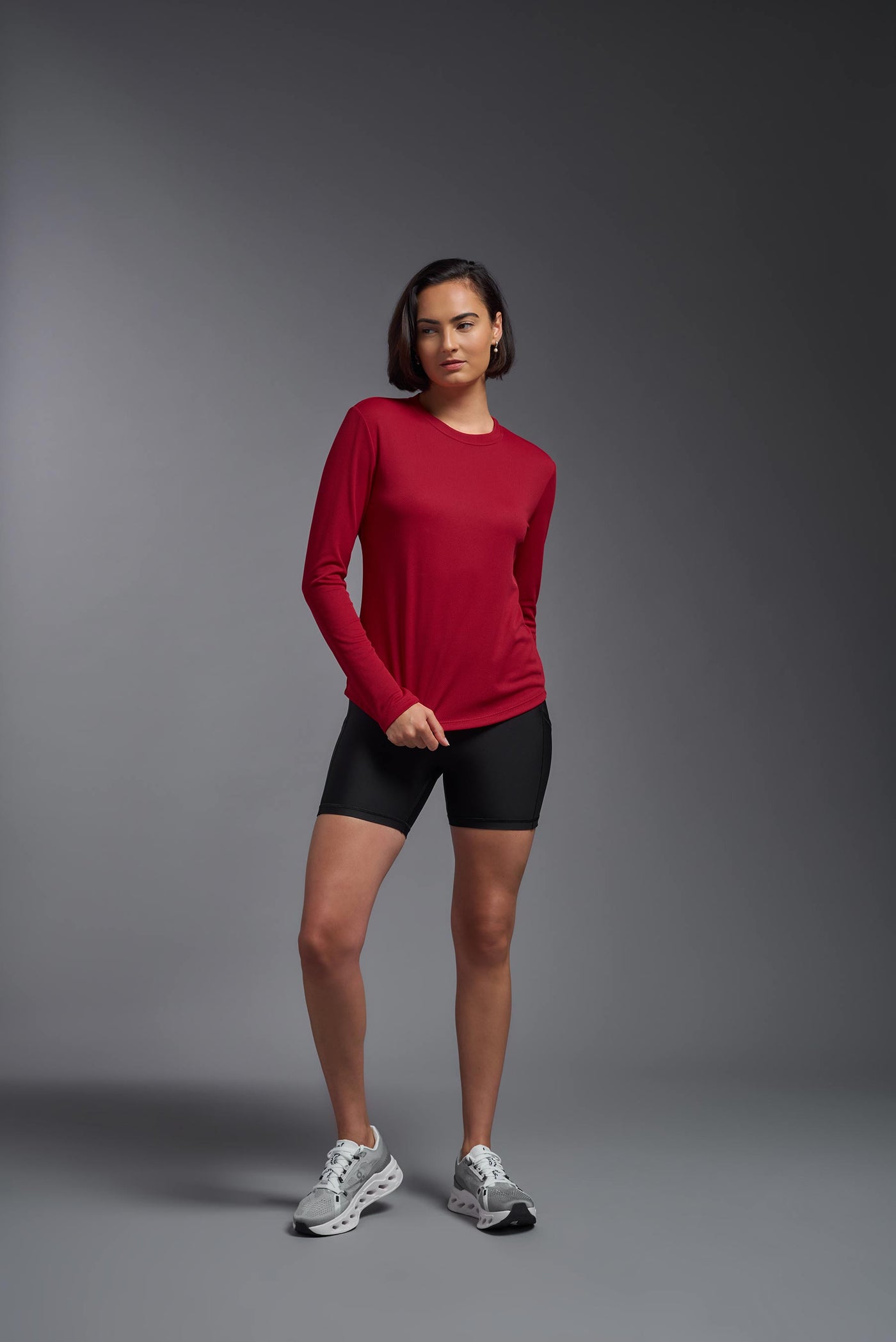 A female model with short brown hair on a gray background, wearing the Women's Oxymesh™ Long Sleeve Tech Tee in the color true red from a full body front view. This shirt is made in the USA and is quick-drying and moisture-wicking. #color_true-red