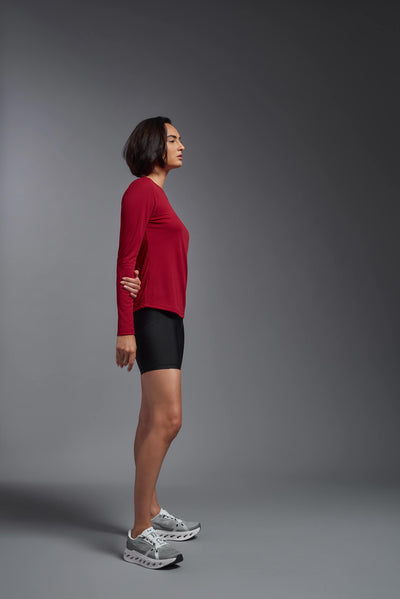 A female model with short brown hair on a gray background, wearing the Women's Oxymesh™ Long Sleeve Tech Tee in the color true red from a full body side view. This shirt is made in the USA and is quick-drying and moisture-wicking. #color_true-red