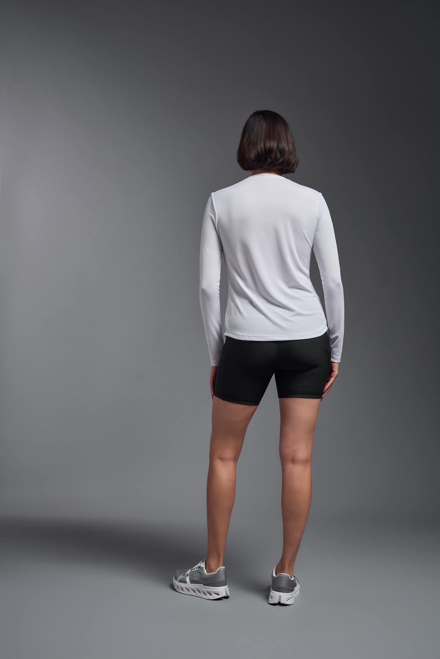 A female model with short brown hair on a gray background, wearing the Women's Oxymesh™ Long Sleeve Tech Tee in the color white from a full body back view. This shirt is made in the USA and is quick-drying and moisture-wicking. #color_white