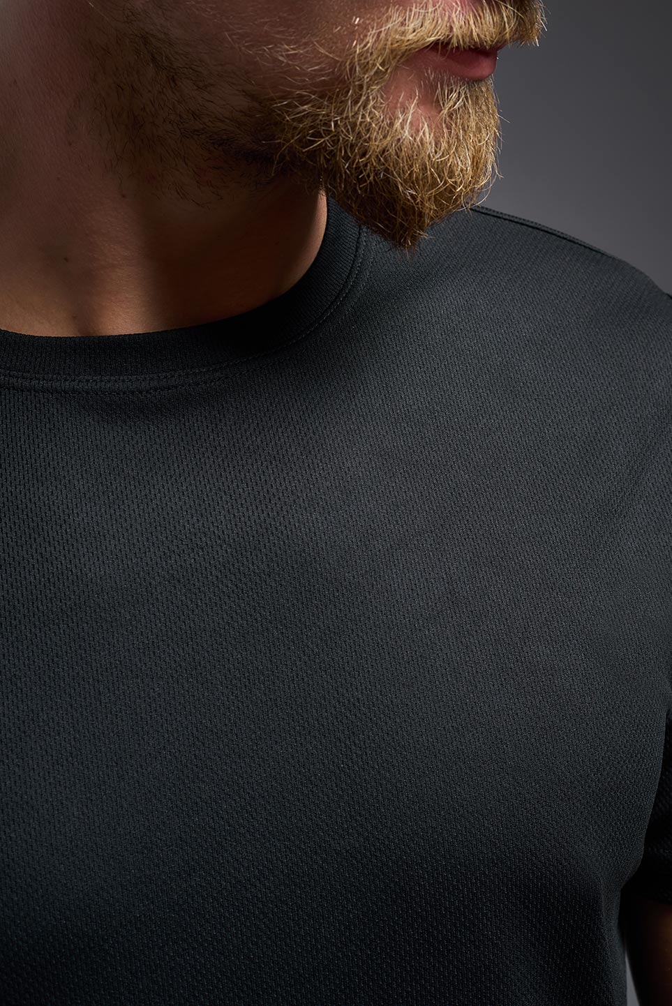 A blonde male model with a beard on a gray background, wearing the Men's Oxymesh™ Crewneck Tech Tee in the color black from a closeup collar front view. This shirt is made in the USA and is quick-drying and moisture-wicking. #color_black