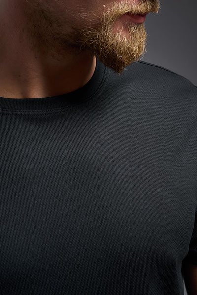 A blonde male model with a beard on a gray background, wearing the Men's Oxymesh™ Crewneck Tech Tee in the color black from a closeup collar front view. This shirt is made in the USA and is quick-drying and moisture-wicking. #color_black