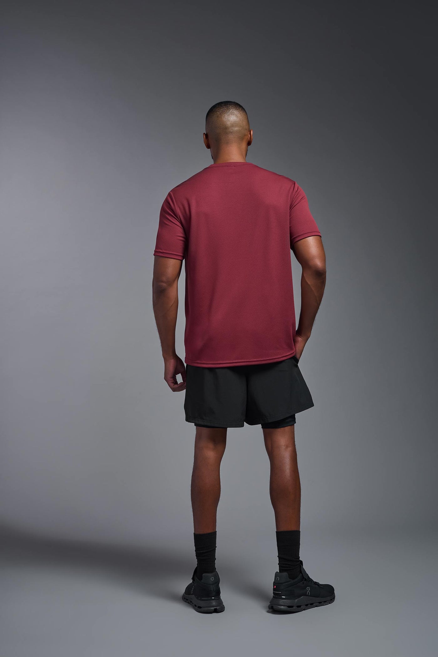 An African American male model with a beard on a gray background, wearing the Men's Oxymesh™ Crewneck Tech Tee in the color cardinal from a full body back view. This shirt is made in the USA and is quick-drying and moisture-wicking. #color_cardinal