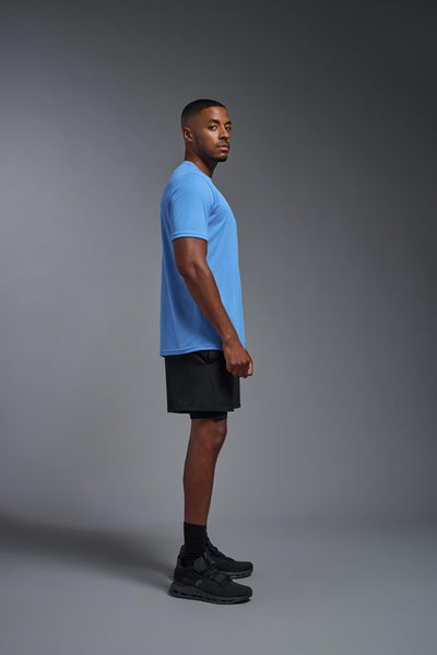 An African American male model with a beard on a gray background, wearing the Men's Oxymesh™ Crewneck Tech Tee in the color carolina blue from a full body side view. This shirt is made in the USA and is quick-drying and moisture-wicking. #color_carolina-blue