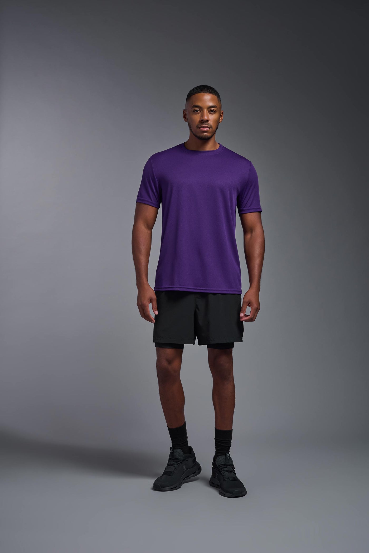 An African American male model with a beard on a gray background, wearing the Men's Oxymesh™ Crewneck Tech Tee in the color dark purple from a full body front view. This shirt is made in the USA and is quick-drying and moisture-wicking. #color_dark-purple