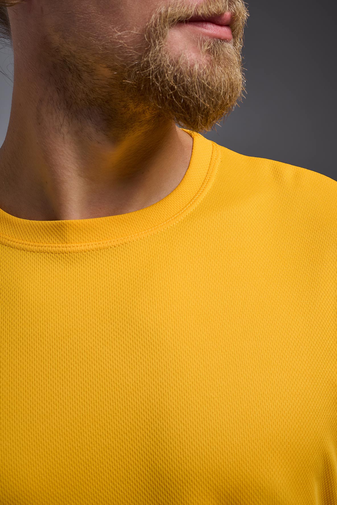 A blonde male model with a beard on a gray background, wearing the Men's Oxymesh™ Crewneck Tech Tee in the color gold from a closeup collar front view. This shirt is made in the USA and is quick-drying and moisture-wicking. #color_gold