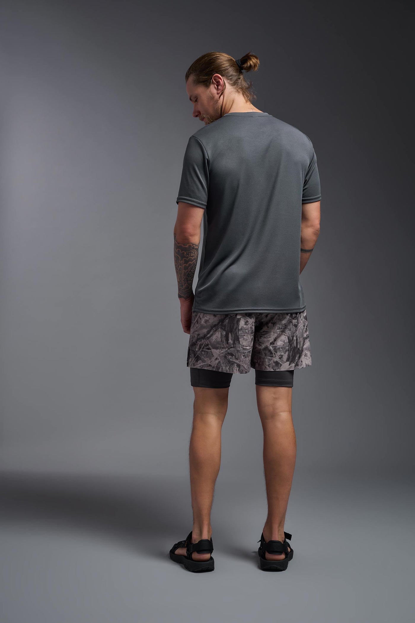 A blonde male model with a beard on a gray background, wearing the Men's Oxymesh™ Crewneck Tech Tee in the color graphite gray from a full body back view. This shirt is made in the USA and is quick-drying and moisture-wicking. #color_graphite-gray