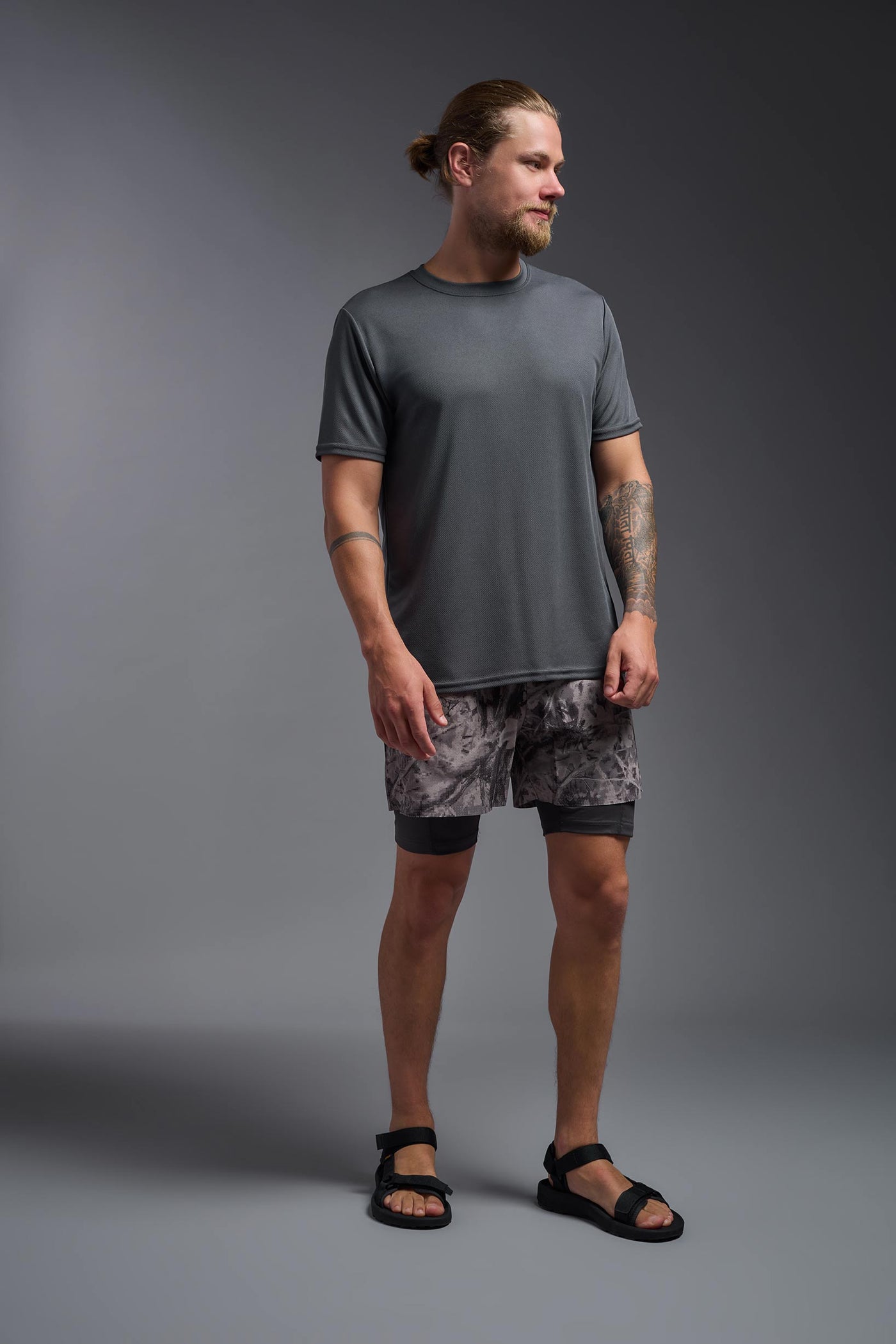 A blonde male model with a beard on a gray background, wearing the Men's Oxymesh™ Crewneck Tech Tee in the color graphite gray from a full body front view. This shirt is made in the USA and is quick-drying and moisture-wicking. #color_graphite-gray