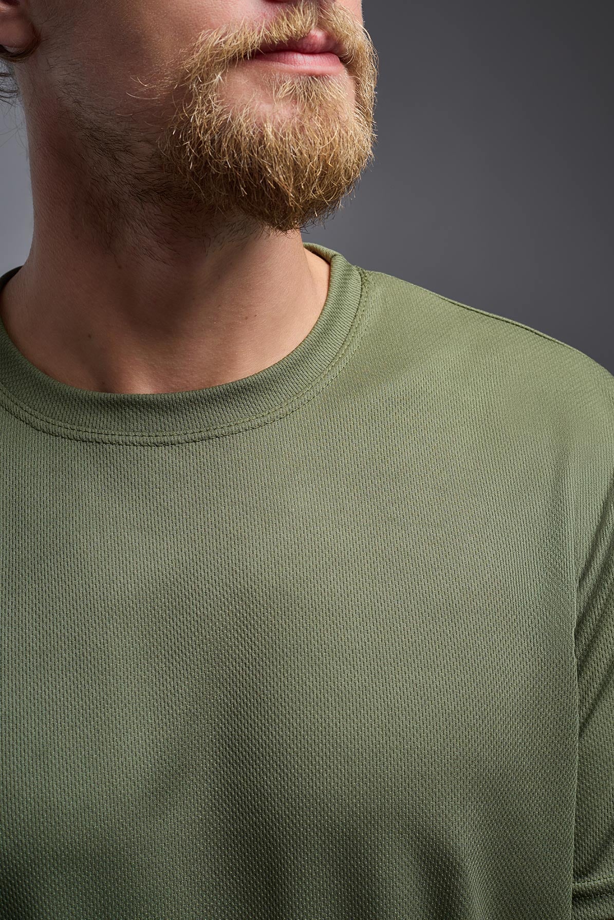 A blonde male model with a beard on a gray background, wearing the Men's Oxymesh™ Crewneck Tech Tee in the color military green from a closeup collar front view. This shirt is made in the USA and is quick-drying and moisture-wicking. #color_military-green