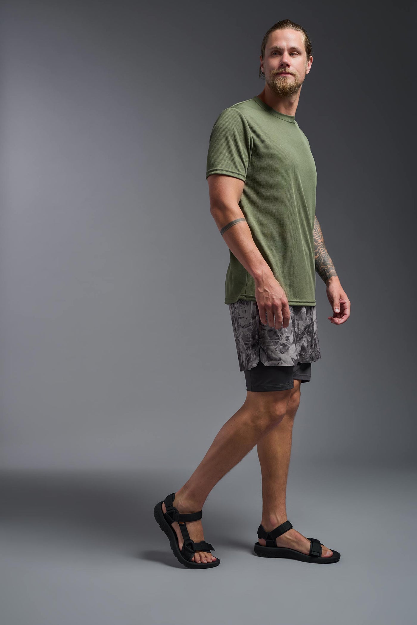 A blonde male model with a beard on a gray background, wearing the Men's Oxymesh™ Crewneck Tech Tee in the color military green from a full body side view. This shirt is made in the USA and is quick-drying and moisture-wicking. #color_military-green