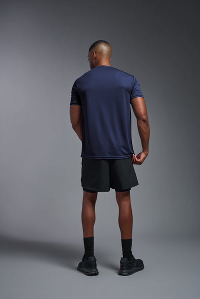 An African American male model with a beard on a gray background, wearing the Men's Oxymesh™ Crewneck Tech Tee in the color navy blue from a full body back view. This shirt is made in the USA and is quick-drying and moisture-wicking. #color_navy-blue