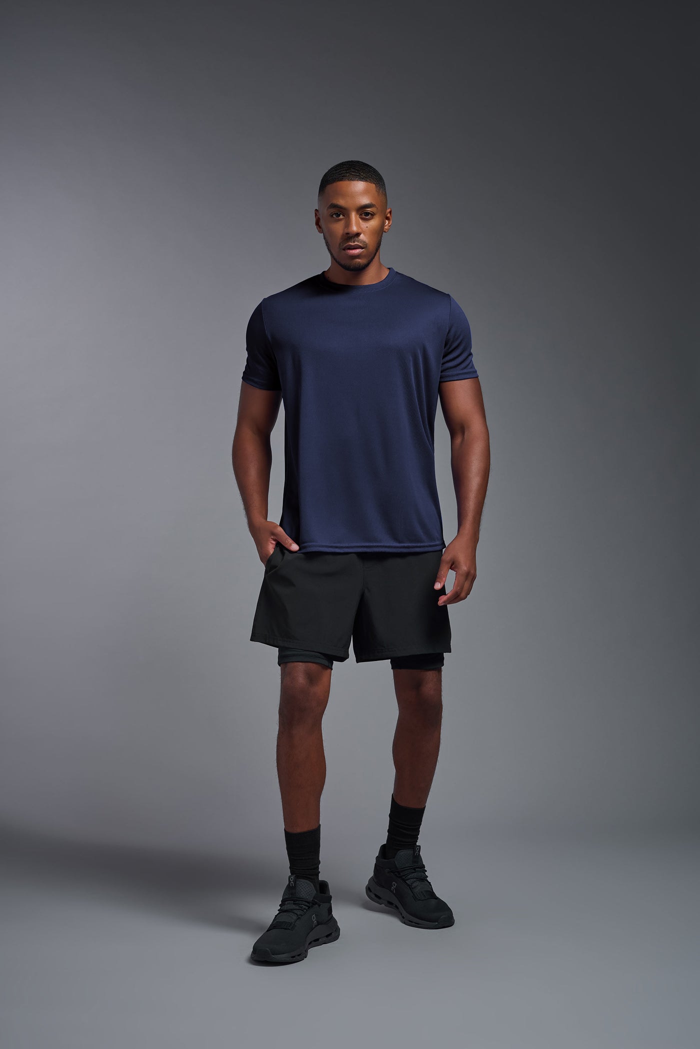 An African American male model with a beard on a gray background, wearing the Men's Oxymesh™ Crewneck Tech Tee in the color navy blue from a full body front view. This shirt is made in the USA and is quick-drying and moisture-wicking. #color_navy-blue