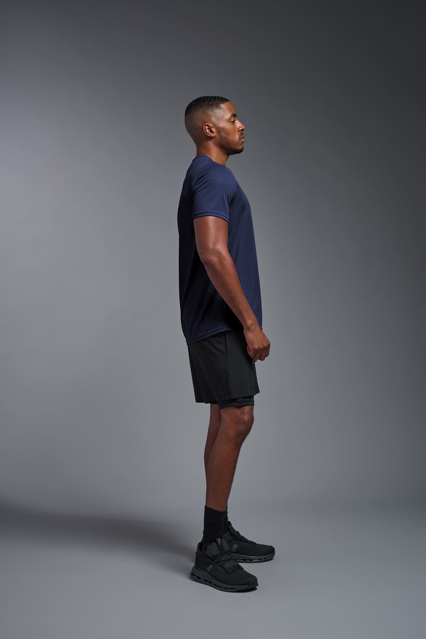 An African American male model with a beard on a gray background, wearing the Men's Oxymesh™ Crewneck Tech Tee in the color navy blue from a full body side view. This shirt is made in the USA and is quick-drying and moisture-wicking. #color_navy-blue