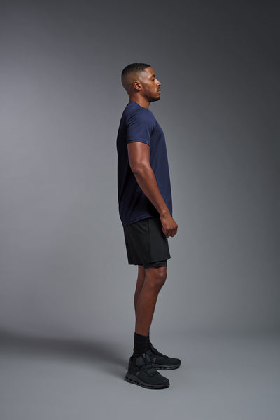 An African American male model with a beard on a gray background, wearing the Men's Oxymesh™ Crewneck Tech Tee in the color navy blue from a full body side view. This shirt is made in the USA and is quick-drying and moisture-wicking. #color_navy-blue