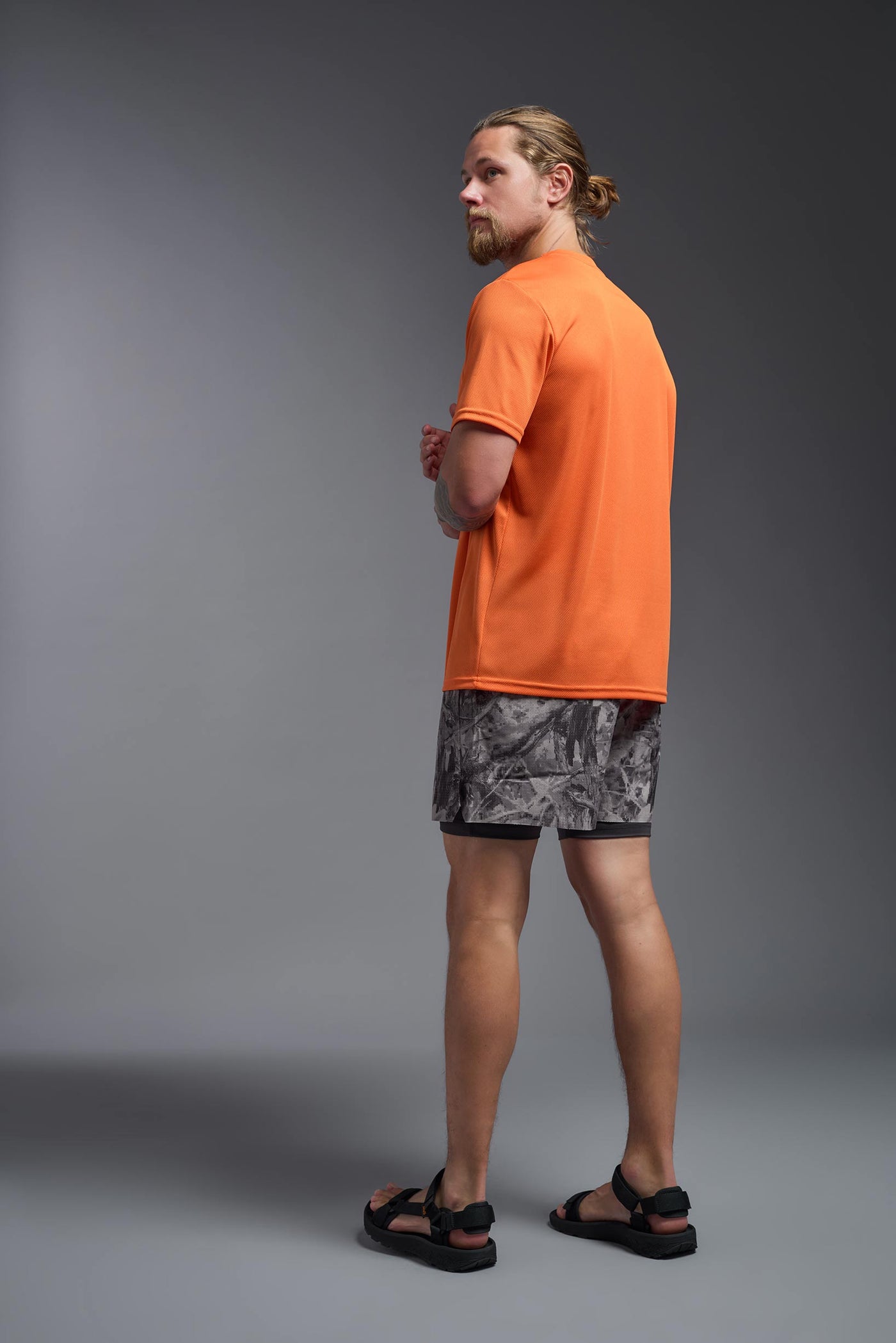 A blonde male model with a beard on a gray background, wearing the Men's Oxymesh™ Crewneck Tech Tee in the color orange from a full body back view. This shirt is made in the USA and is quick-drying and moisture-wicking. #color_orange
