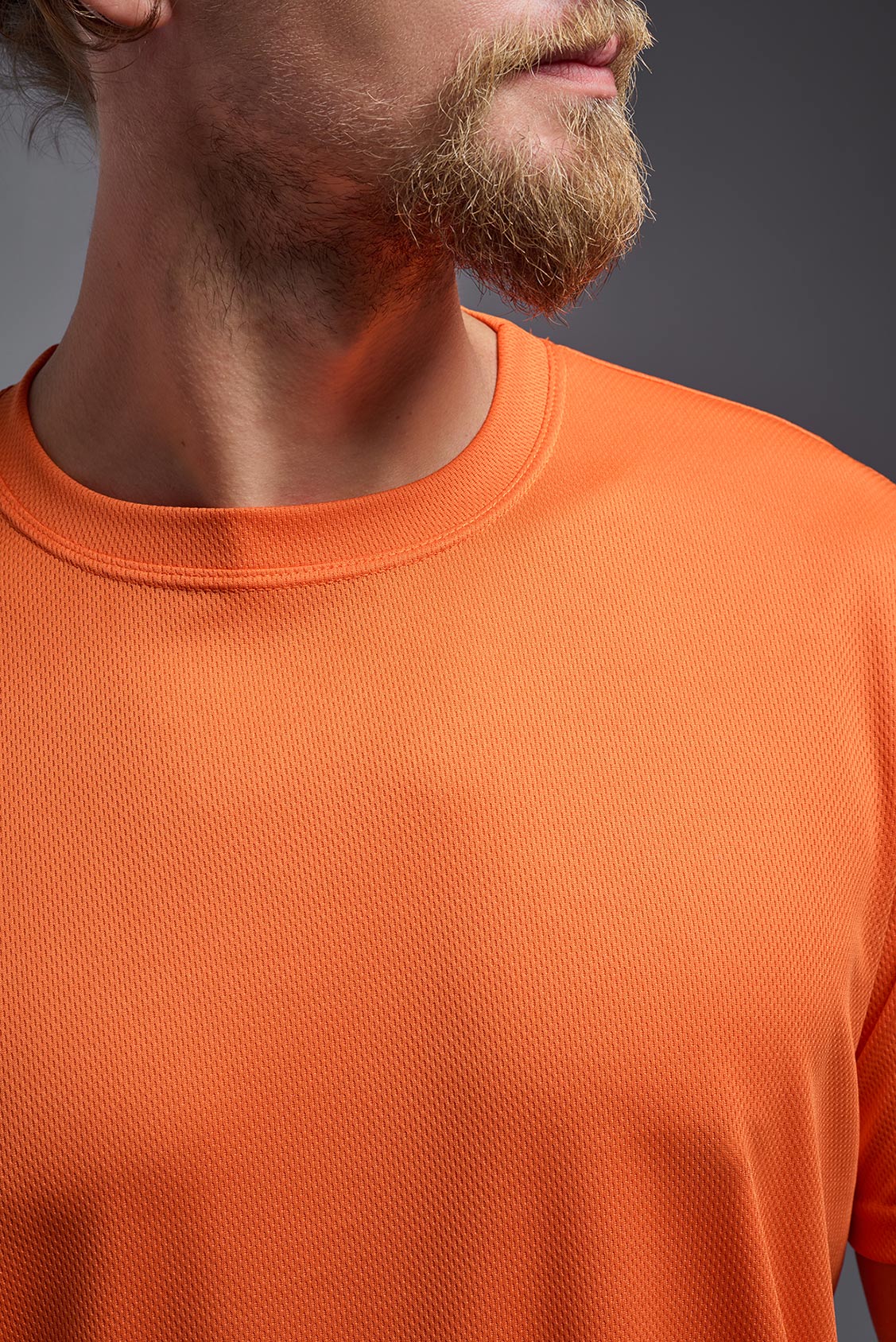 A blonde male model with a beard on a gray background, wearing the Men's Oxymesh™ Crewneck Tech Tee in the color orange from a closeup collar front view. This shirt is made in the USA and is quick-drying and moisture-wicking. #color_orange