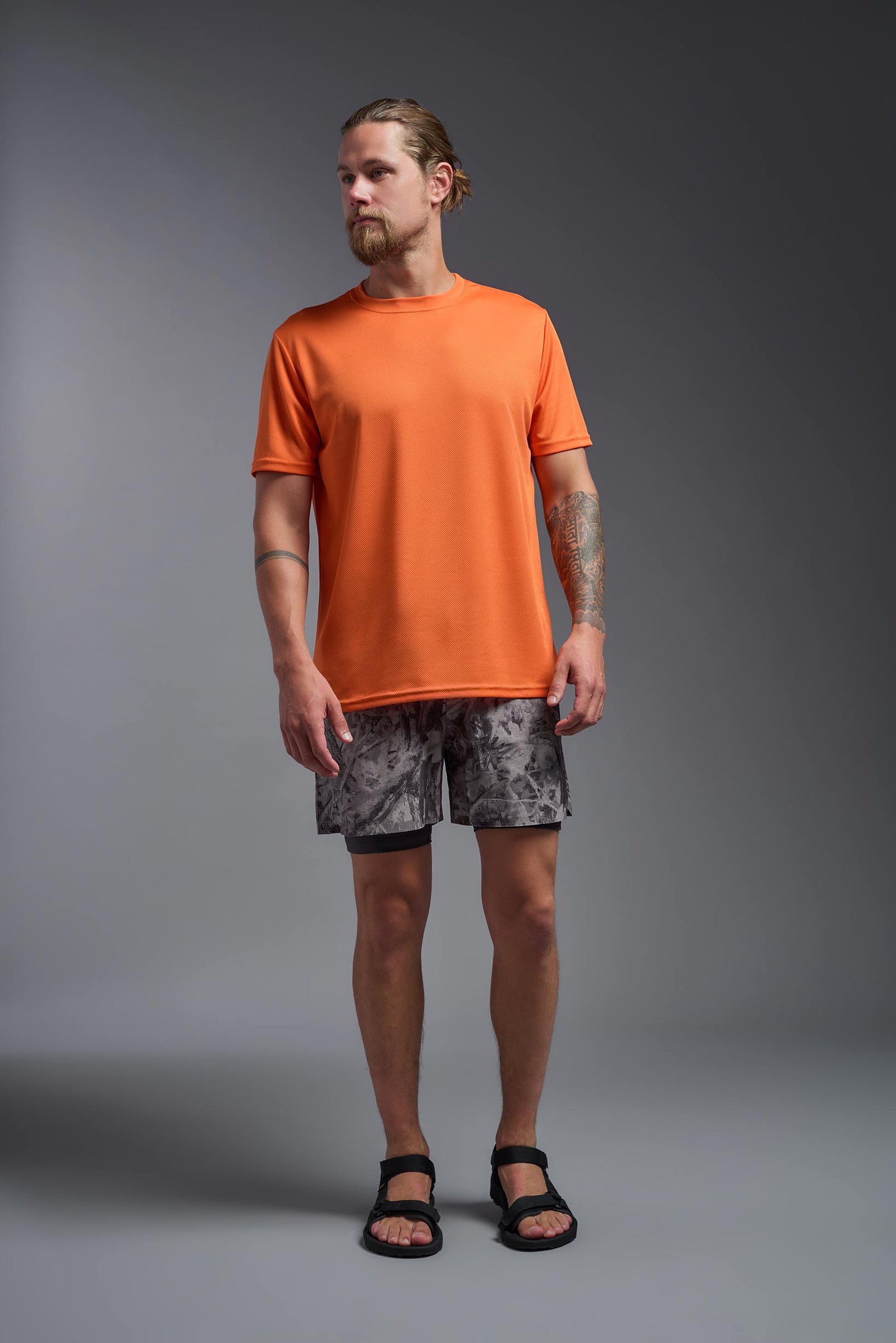 A blonde male model with a beard on a gray background, wearing the Men's Oxymesh™ Crewneck Tech Tee in the color orange from a full body front view. This shirt is made in the USA and is quick-drying and moisture-wicking. #color_orange