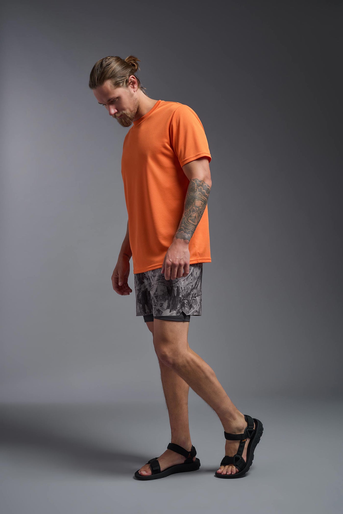 A blonde male model with a beard on a gray background, wearing the Men's Oxymesh™ Crewneck Tech Tee in the color orange from a full body side view. This shirt is made in the USA and is quick-drying and moisture-wicking. #color_orange