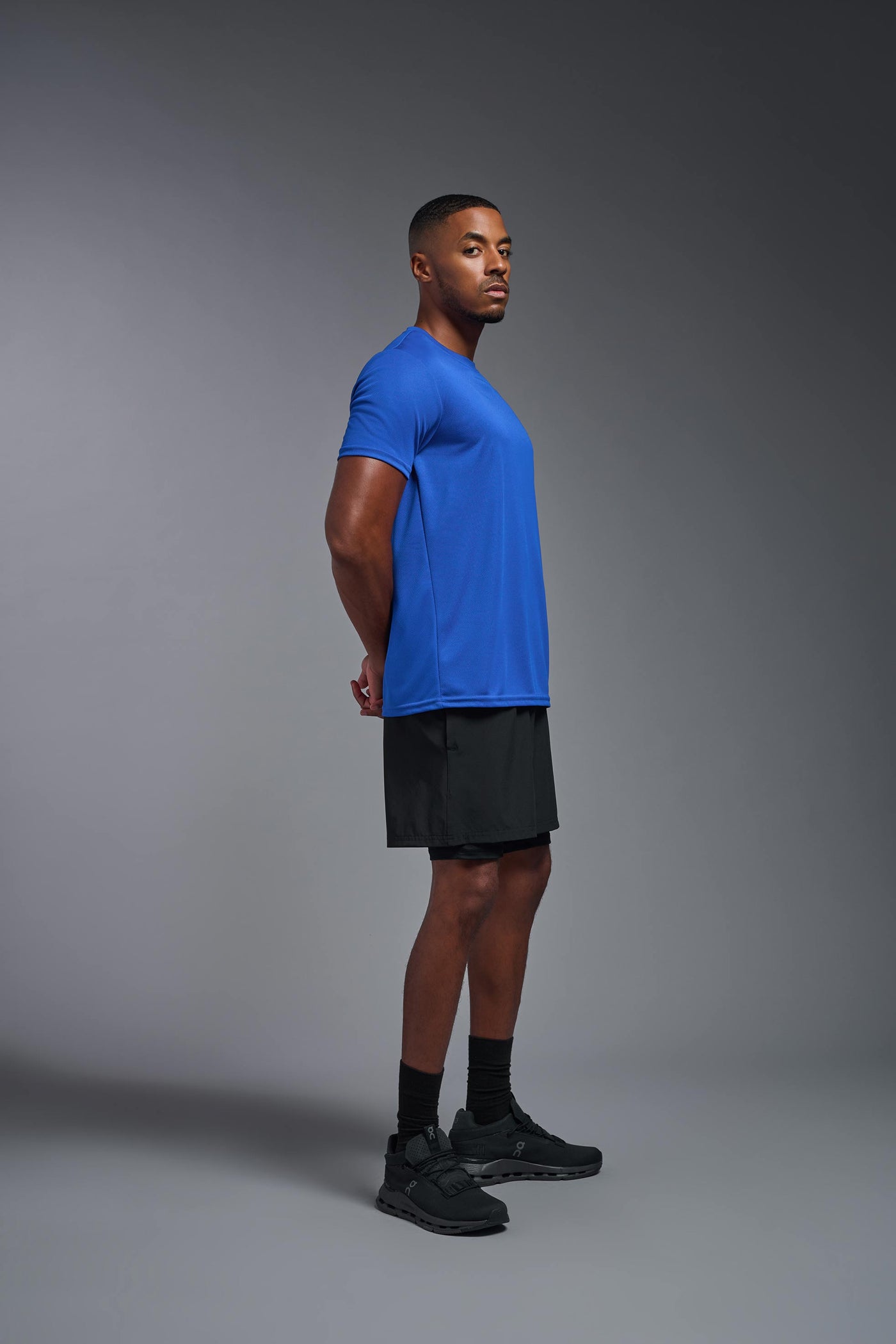 An African American male model with a beard on a gray background, wearing the Men's Oxymesh™ Crewneck Tech Tee in the color royal blue from a full body side view. This shirt is made in the USA and is quick-drying and moisture-wicking. #color_royal-blue
