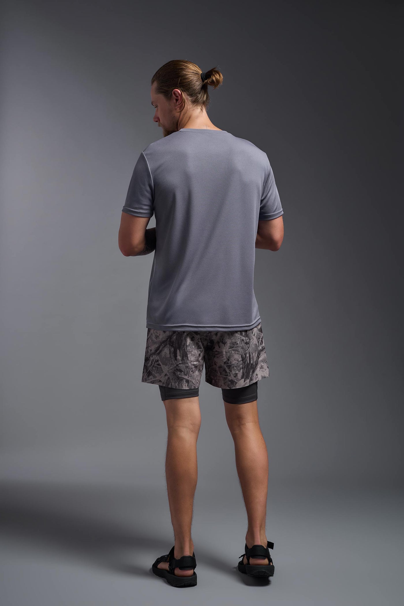 A blonde male model with a beard on a gray background, wearing the Men's Oxymesh™ Crewneck Tech Tee in the color steel gray from a full body back view. This shirt is made in the USA and is quick-drying and moisture-wicking. #color_steel-gray